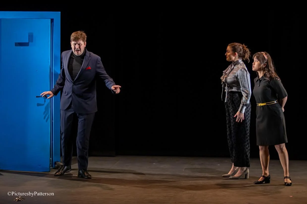 Three people on a stage, two women standing side by side and a man appearing to speak or argue, with a large blue door in the background.