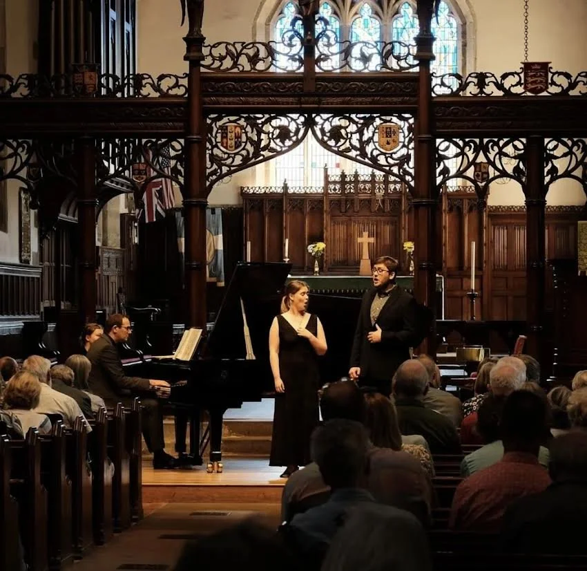Two performers, a man and a woman, stand on stage in front of a grand piano during a concert inside a church or cathedral with stained glass windows and wooden decor.