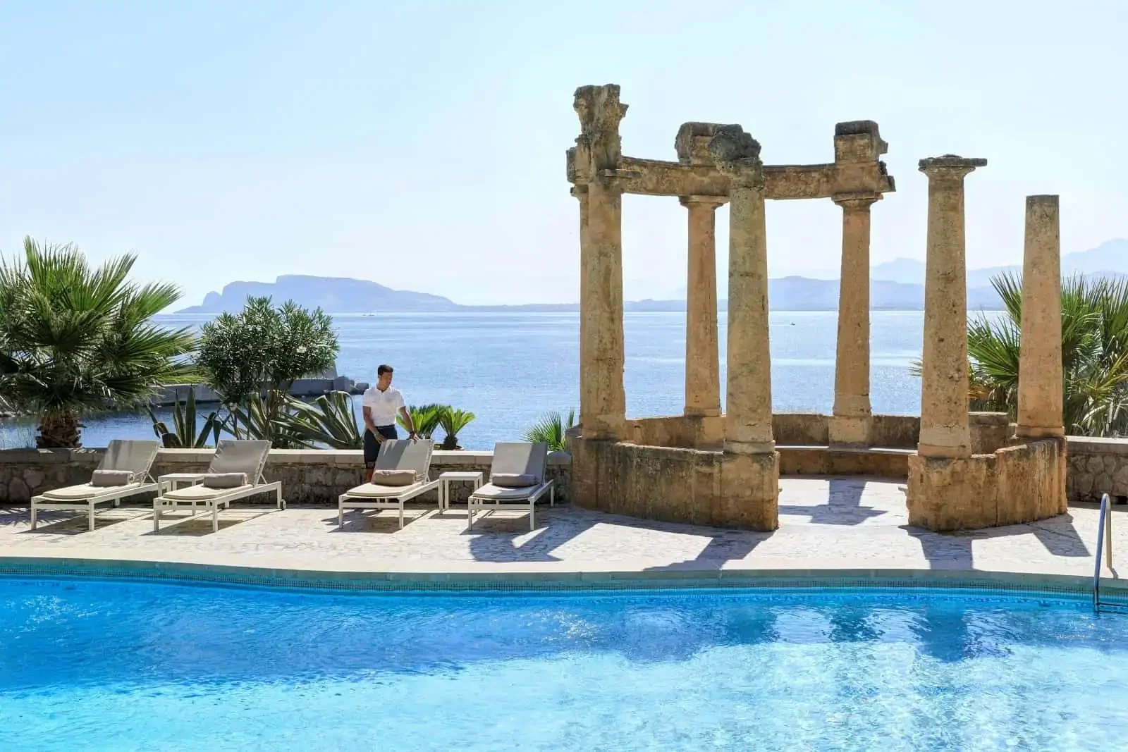 Outdoor pool area with lounge chairs, ancient ruins, palm trees, and a view of the sea and distant islands.