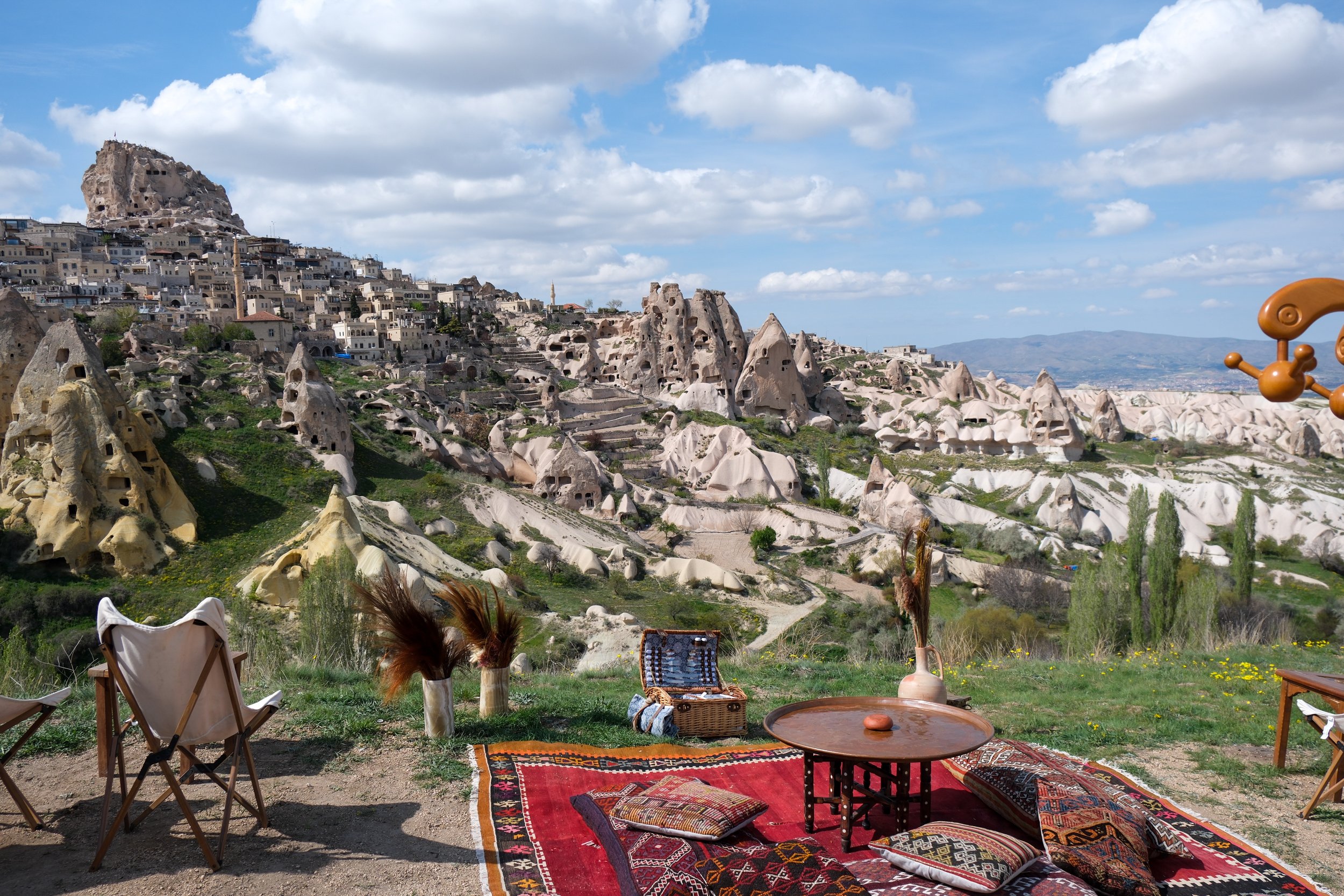 A scenic outdoor view of Cappadocia, Turkey, with rock formations, cave dwellings, and a hillside town in the background, viewed from a decorated sitting area with chairs, carpets, pillows, and vases in the foreground.