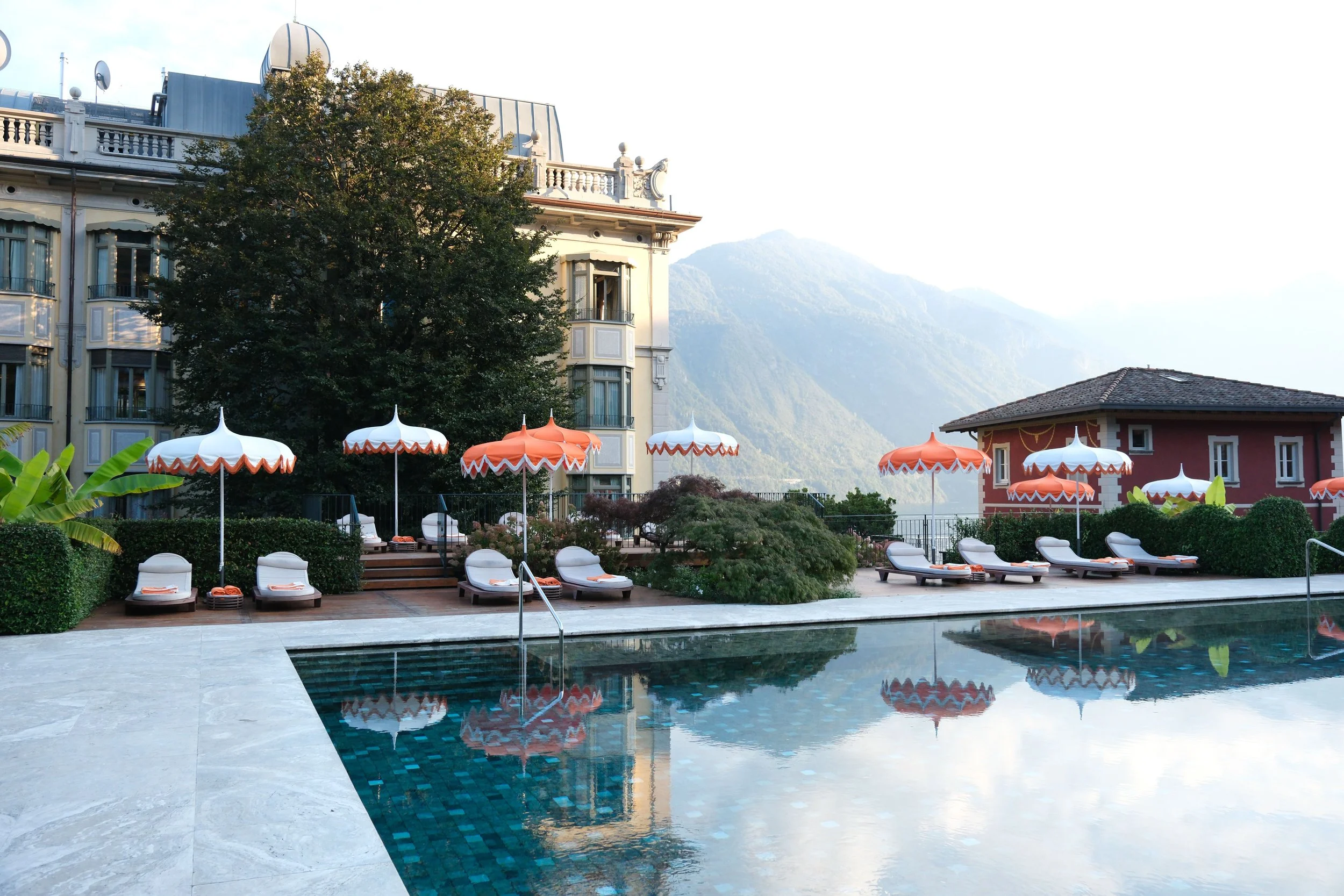 Poolside lounge chairs with orange and white umbrellas at a resort, with a large tree, a building, and mountains in the background.