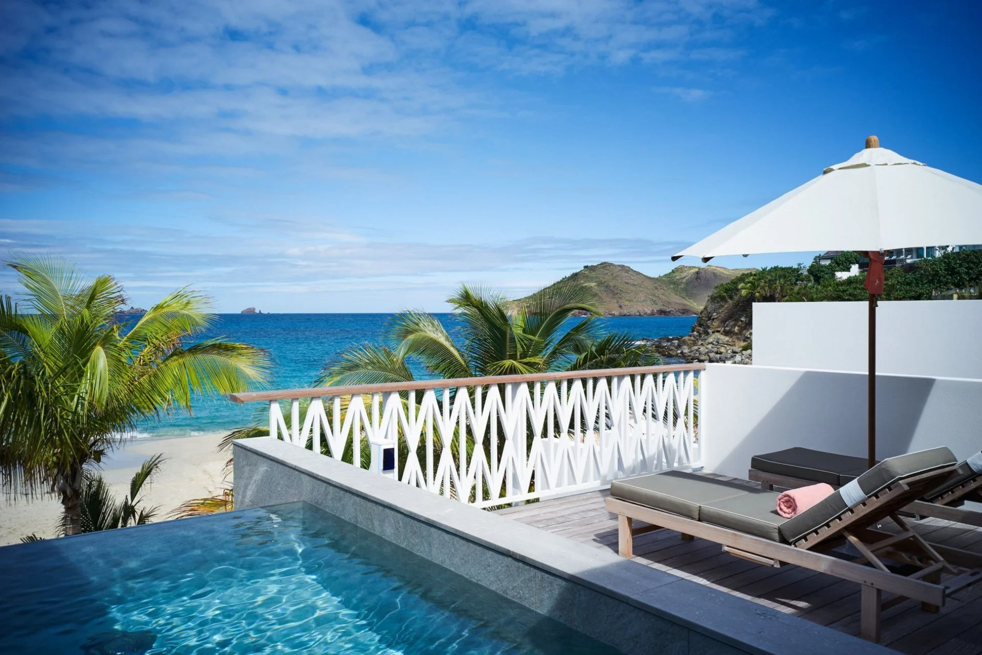 Beachside view with palm trees, ocean, and rocky hills, seen from a balcony with a white railing, lounge chairs, and a white umbrella