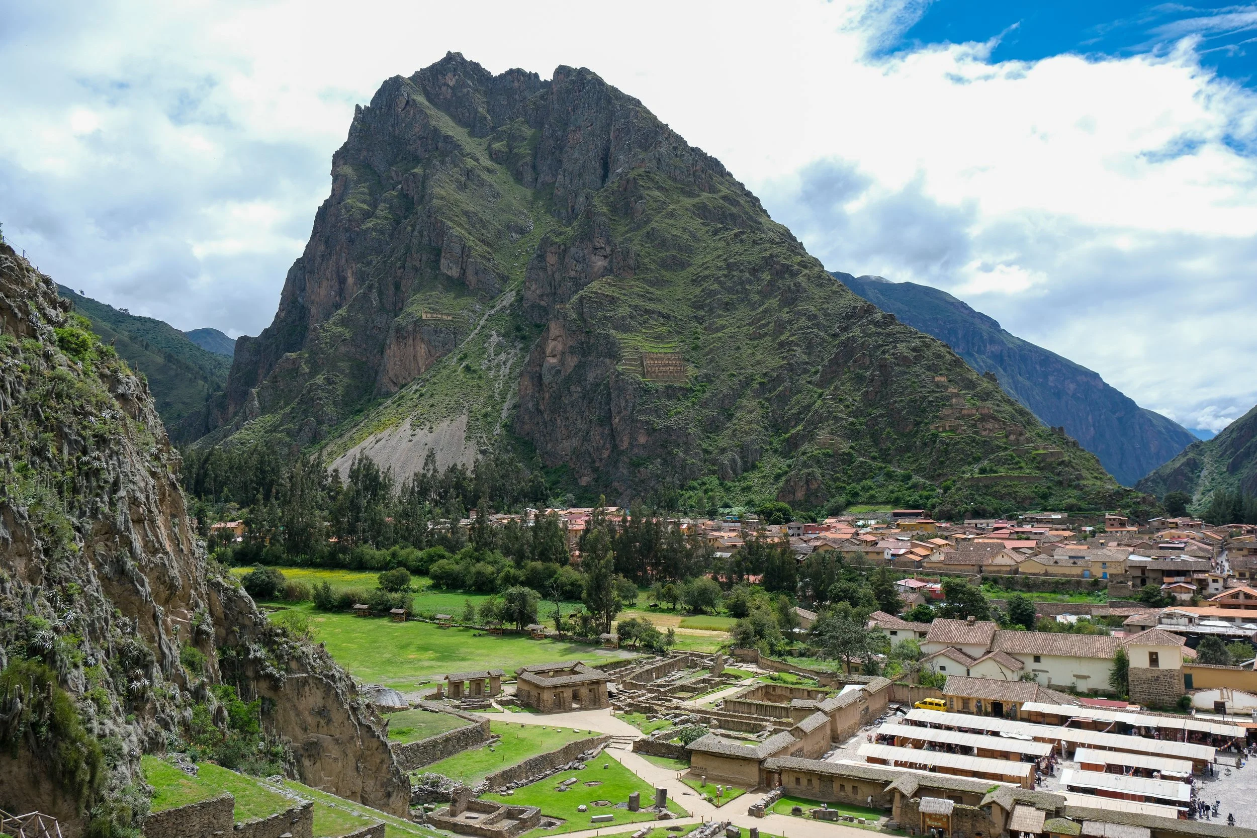 View of Machu Picchu ruins with green terraces, buildings, and pathways in the foreground, surrounded by lush green mountains and a cloudy sky.
