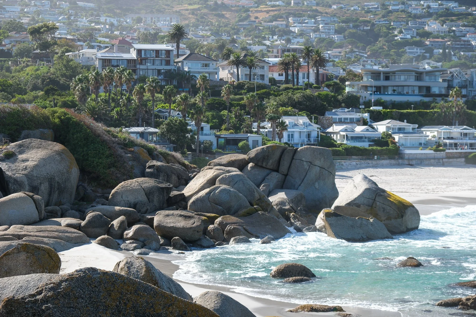 Coastal view with large rocks in the foreground, sandy beach, and modern houses on a hillside surrounded by palm trees in the background.
