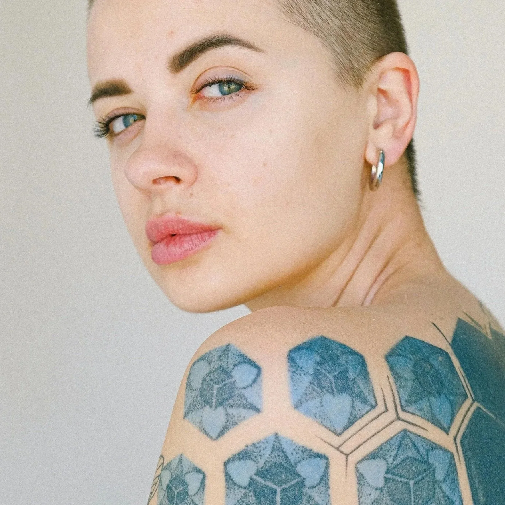 Close-up of a woman with clear skin,  blue eyes, short hair, wearing a hoop earring, and a geometric tattoo on her shoulder.