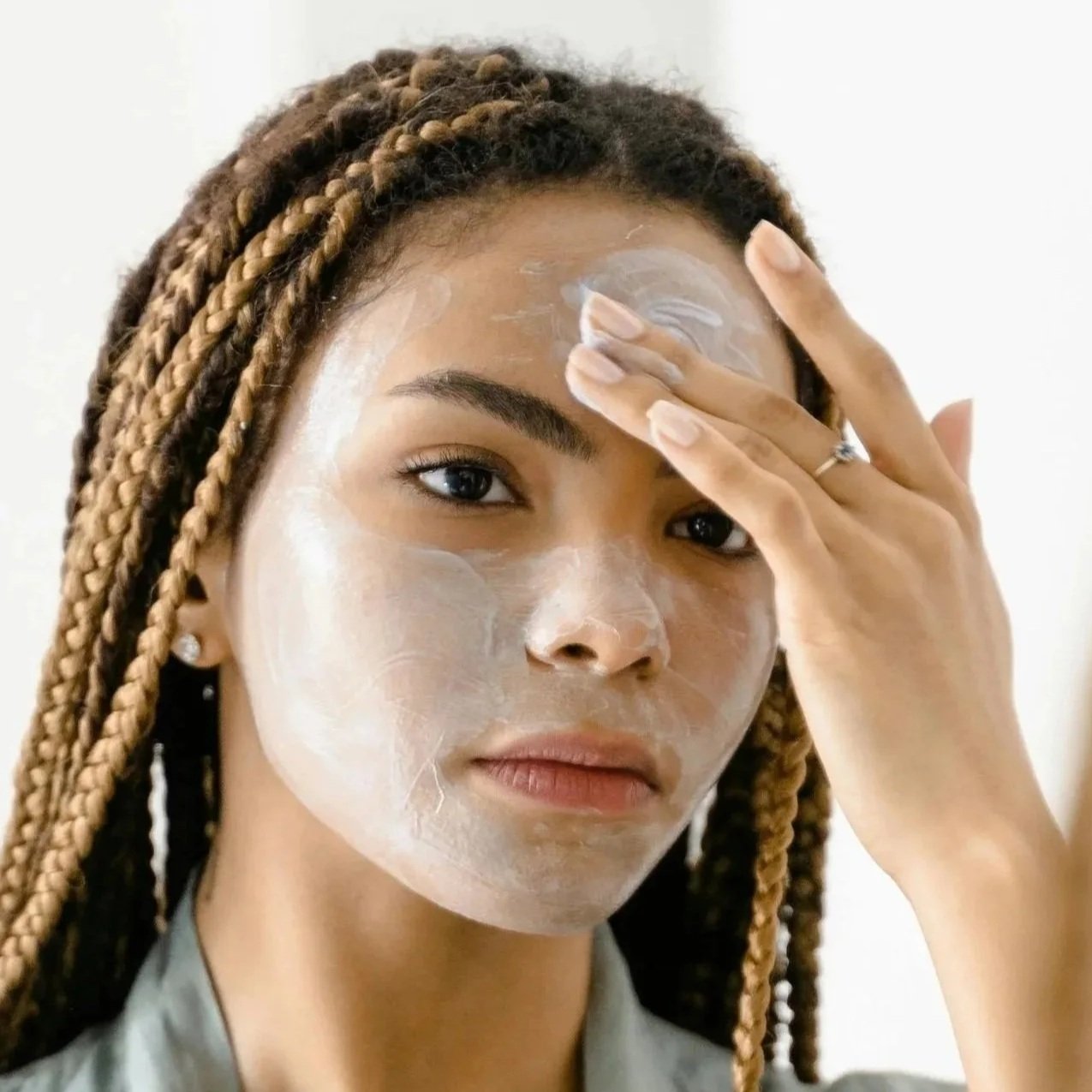 A woman with braided hair applies a white facial mask to her face, touching her forehead.