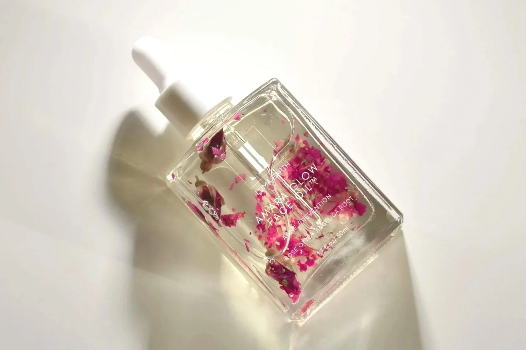 A clear facial oil bottle with a white dropper cap, filled with water and pink flower petals, casting a shadow on a white surface.