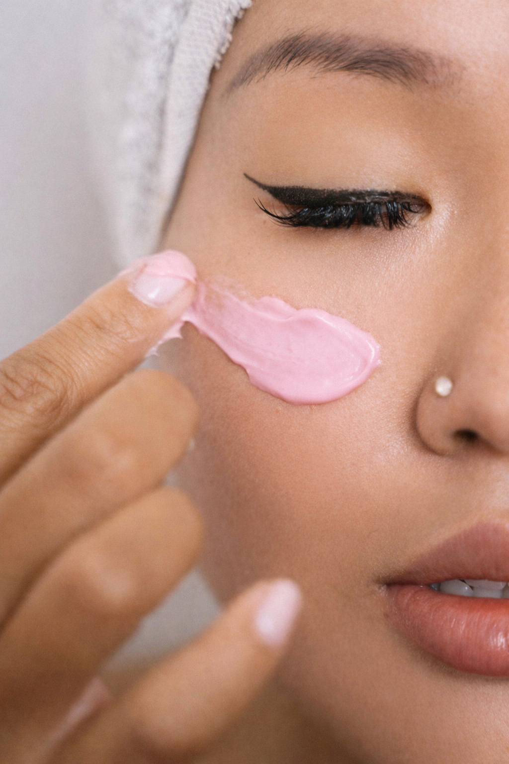 A woman applying pink facial cream to her cheek using her finger, with a towel wrapped around her head.