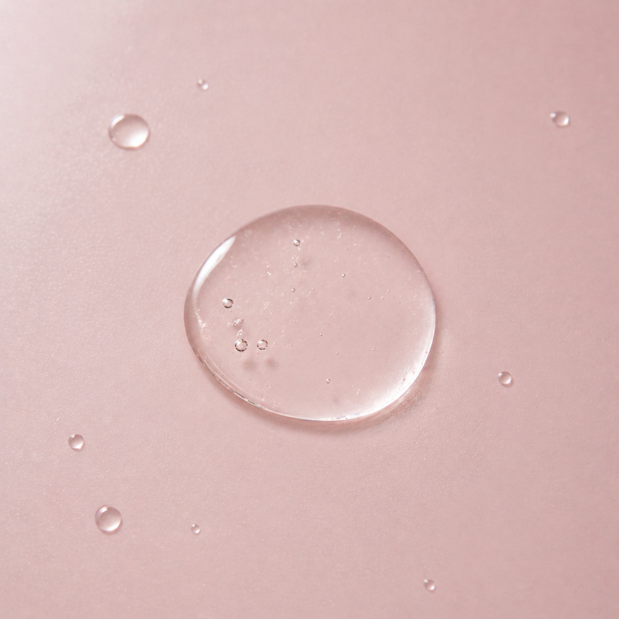 Close-up of a clear water droplet on a pink surface with smaller droplets around it.