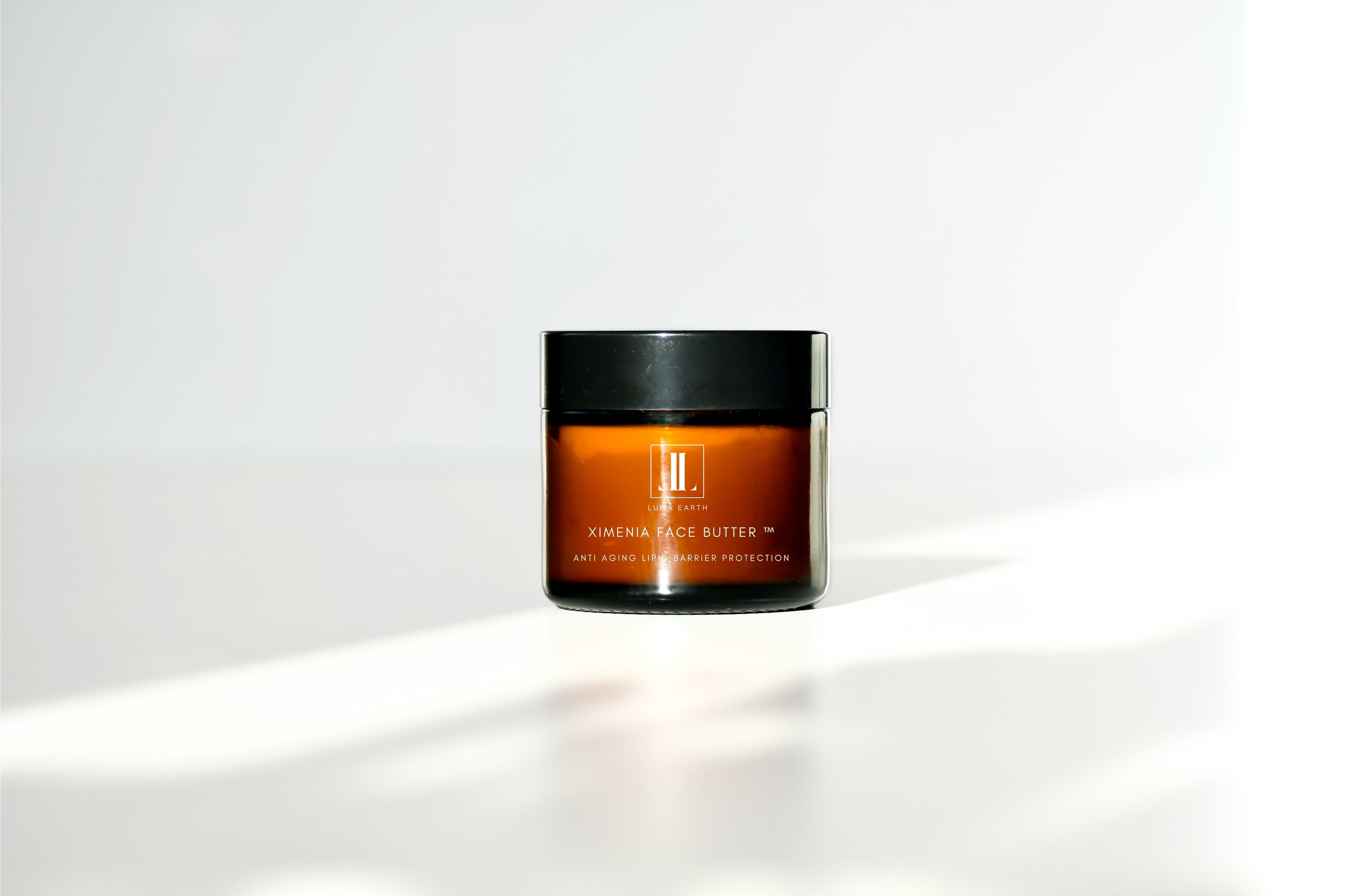 A small brown jar of Lumira Earth Ximenia Face Butter with a black lid, placed on a white surface against a white background.