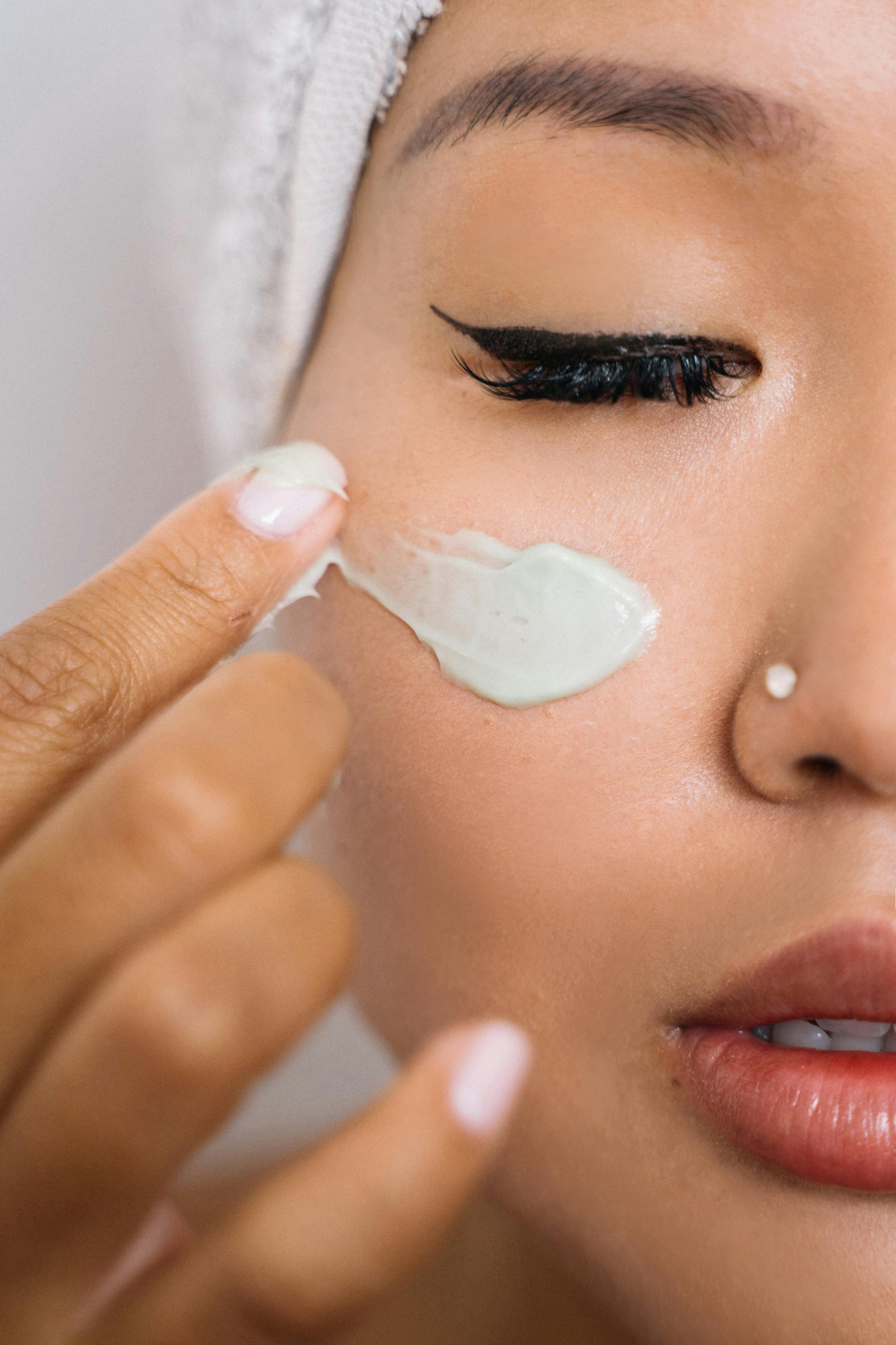 Close-up of a woman with makeup, applying a white cream or mask to her cheek using her finger.