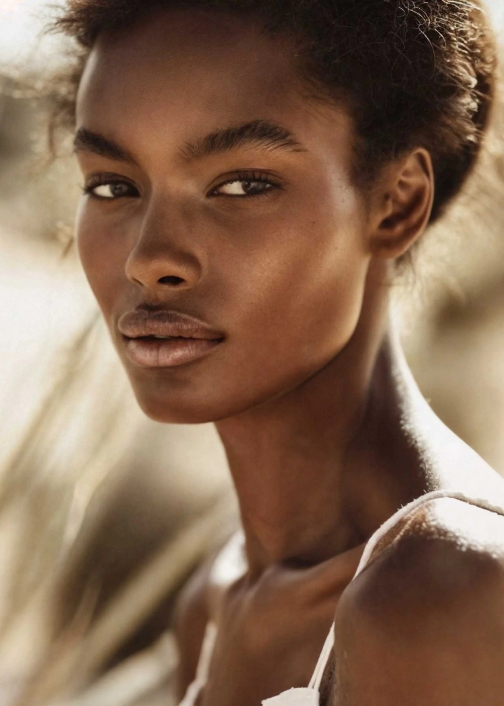 A close-up of a woman with medium brown skin and short curly hair, looking thoughtfully into the camera with sunlight highlighting her face.