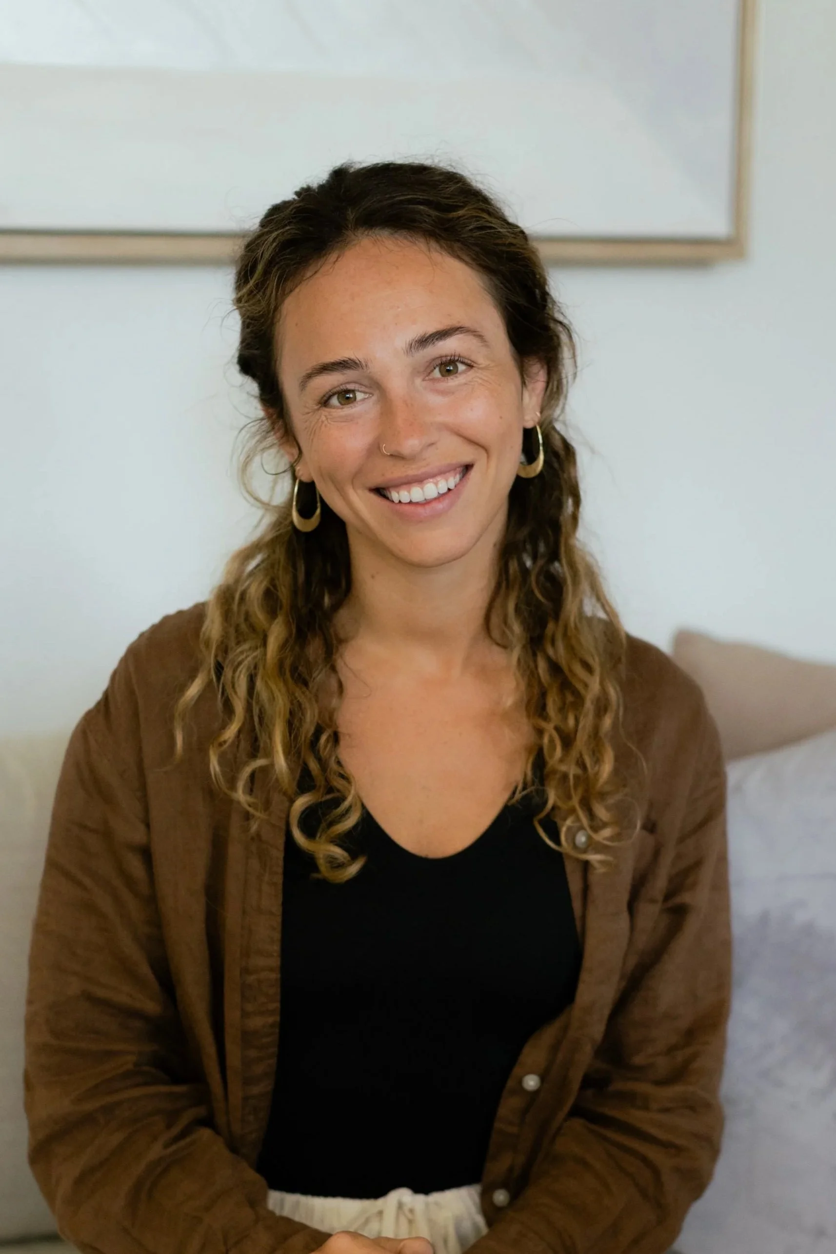 A woman with curly hair smiling at the camera, wearing a black top and a brown jacket, with a white pillow and a beige wall behind her.