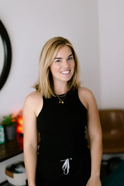 A young woman with blonde hair wearing a black sleeveless top and gold necklaces, smiling in a modern room with a bedroom in the background.