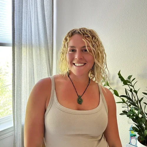 Woman with blonde curly hair smiling in a light-colored tank top, standing next to a potted plant and near a window with blinds in a bright room.