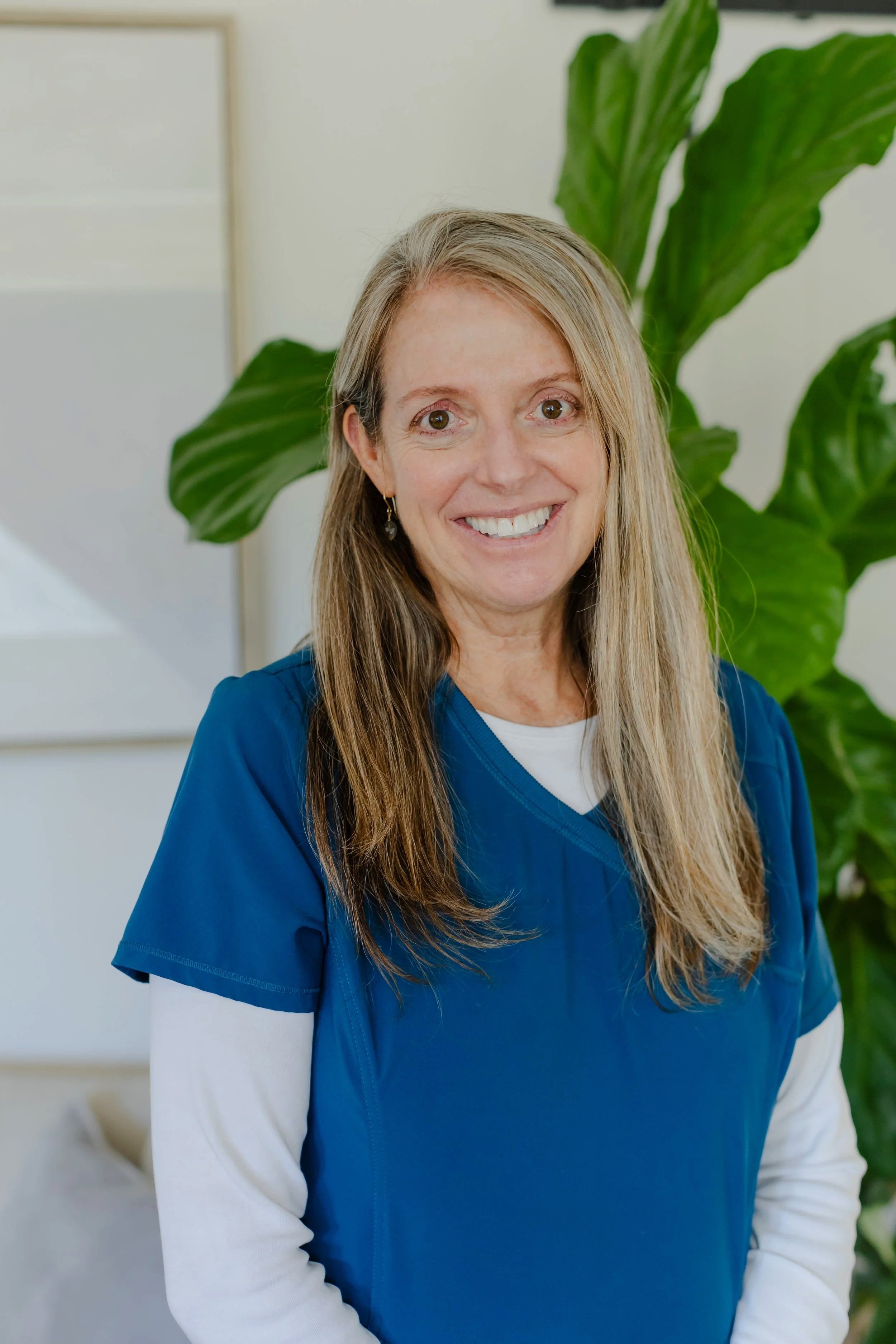 A woman with long, light brown hair wearing blue scrubs, smiling indoors with a large green plant in the background.