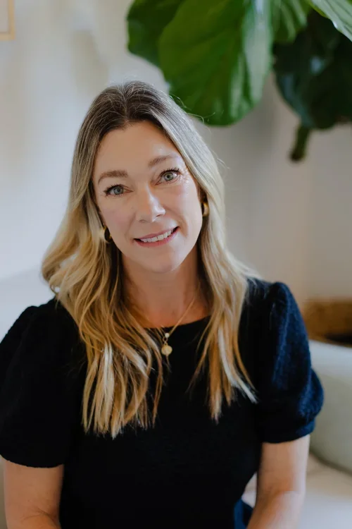A woman with blonde hair and blue eyes smiling while sitting indoors, with large green leaves in the background.