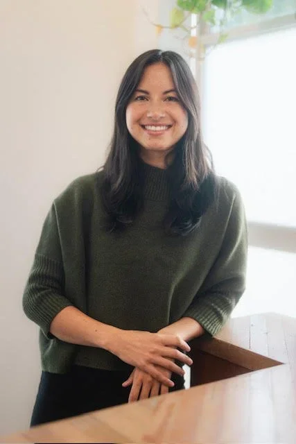 A woman with long dark hair, wearing a dark green sweater, smiling and standing indoors near a window with natural light and some green plants in the background.