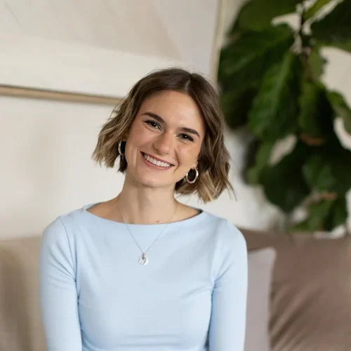 A smiling young woman with short wavy hair wearing a light blue shirt, hoop earrings, and a necklace, sitting indoors with a large green plant in the background.