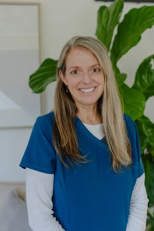 A smiling woman with long blonde hair wearing blue medical scrubs standing in front of a green plant.