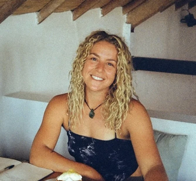 Woman with blonde curly hair smiling in a light-colored tank top, standing next to a potted plant and near a window with blinds in a bright room.
