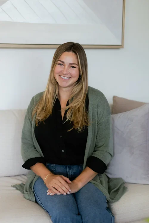 A smiling woman with long blonde hair sitting on a white couch with beige pillows, wearing a black shirt and a green cardigan.