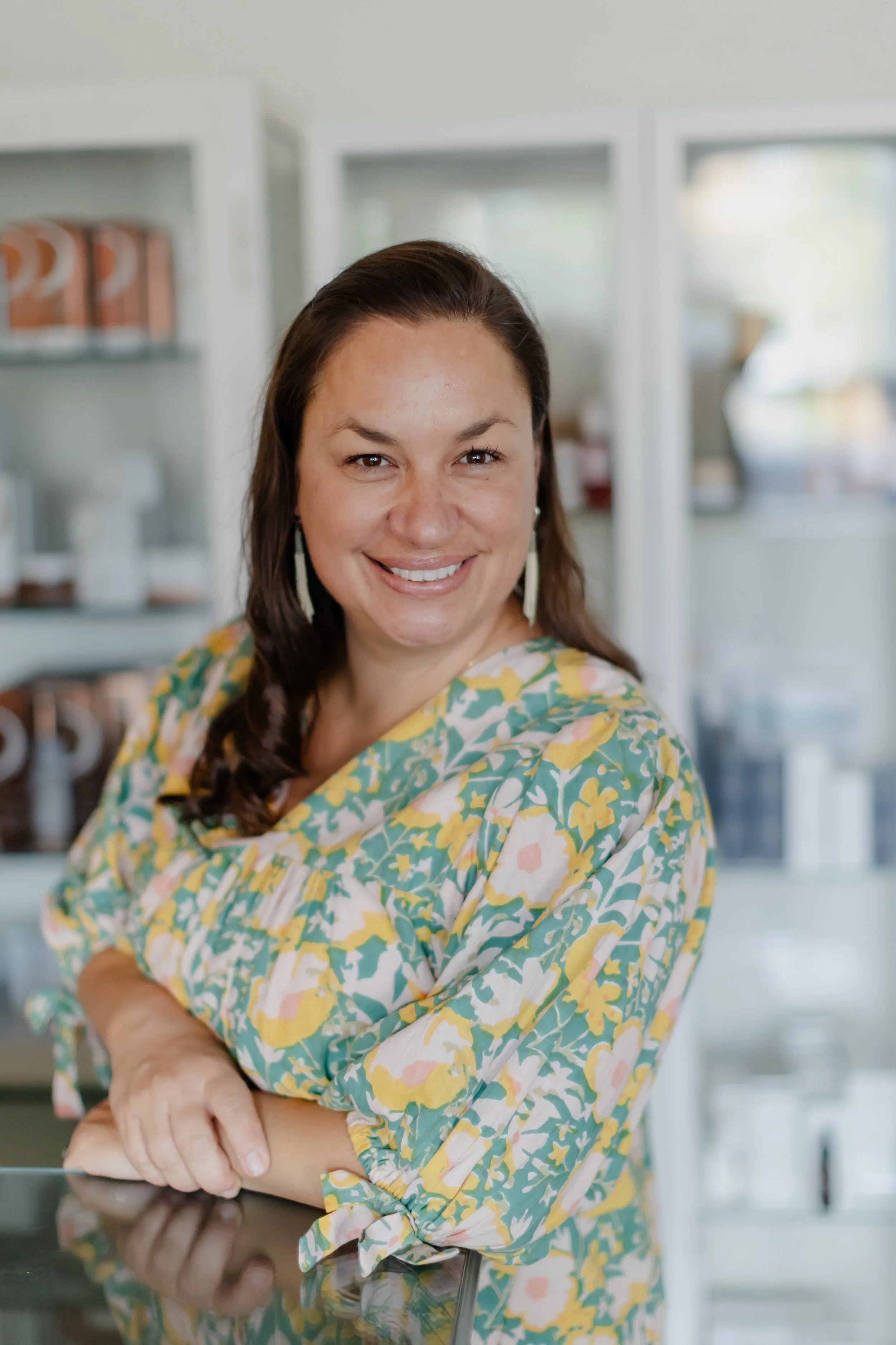 A woman with brown hair wearing a floral dress, smiling, with shelves of containers in the background.