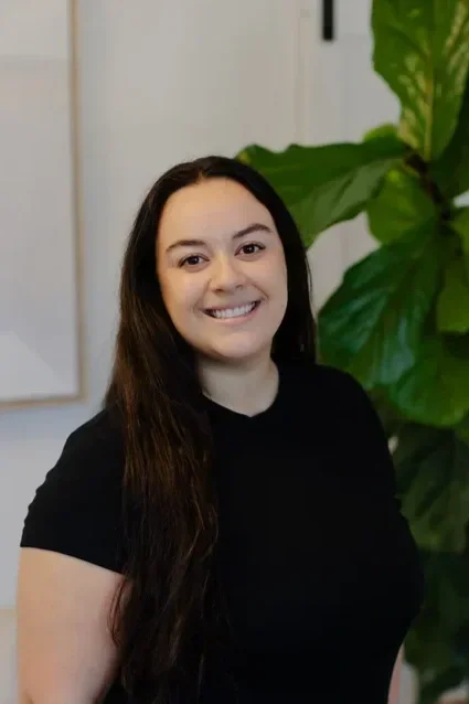 A young woman with long dark hair smiling in front of large green plants indoors.