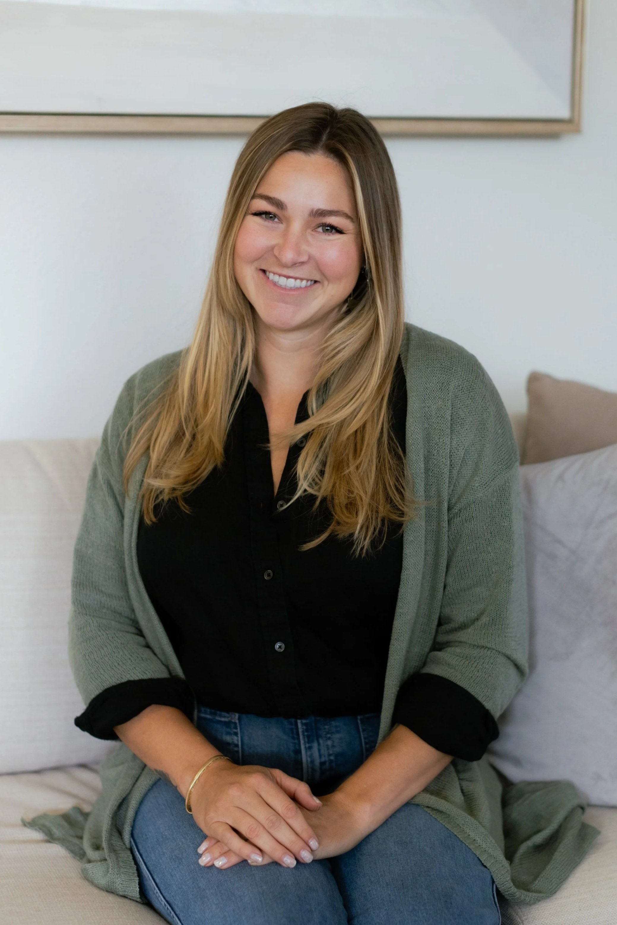 A young woman with long blonde hair smiling, wearing a black shirt and a gray cardigan, sitting on a bed in a bedroom.