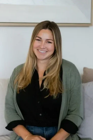 A young woman with long blonde hair smiling, wearing a black shirt and a gray cardigan, sitting on a bed in a bedroom.