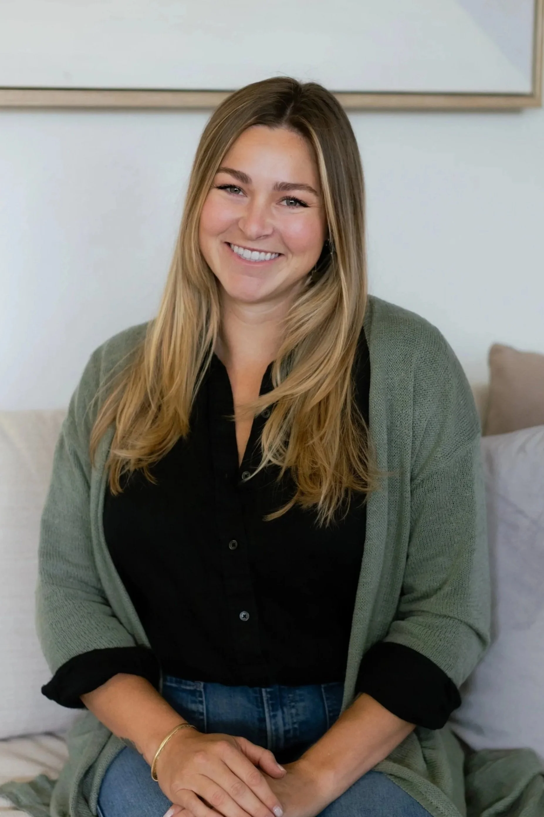 Smiling woman with long, wavy blonde hair wearing a black shirt and gray cardigan, sitting on a couch in a home setting.