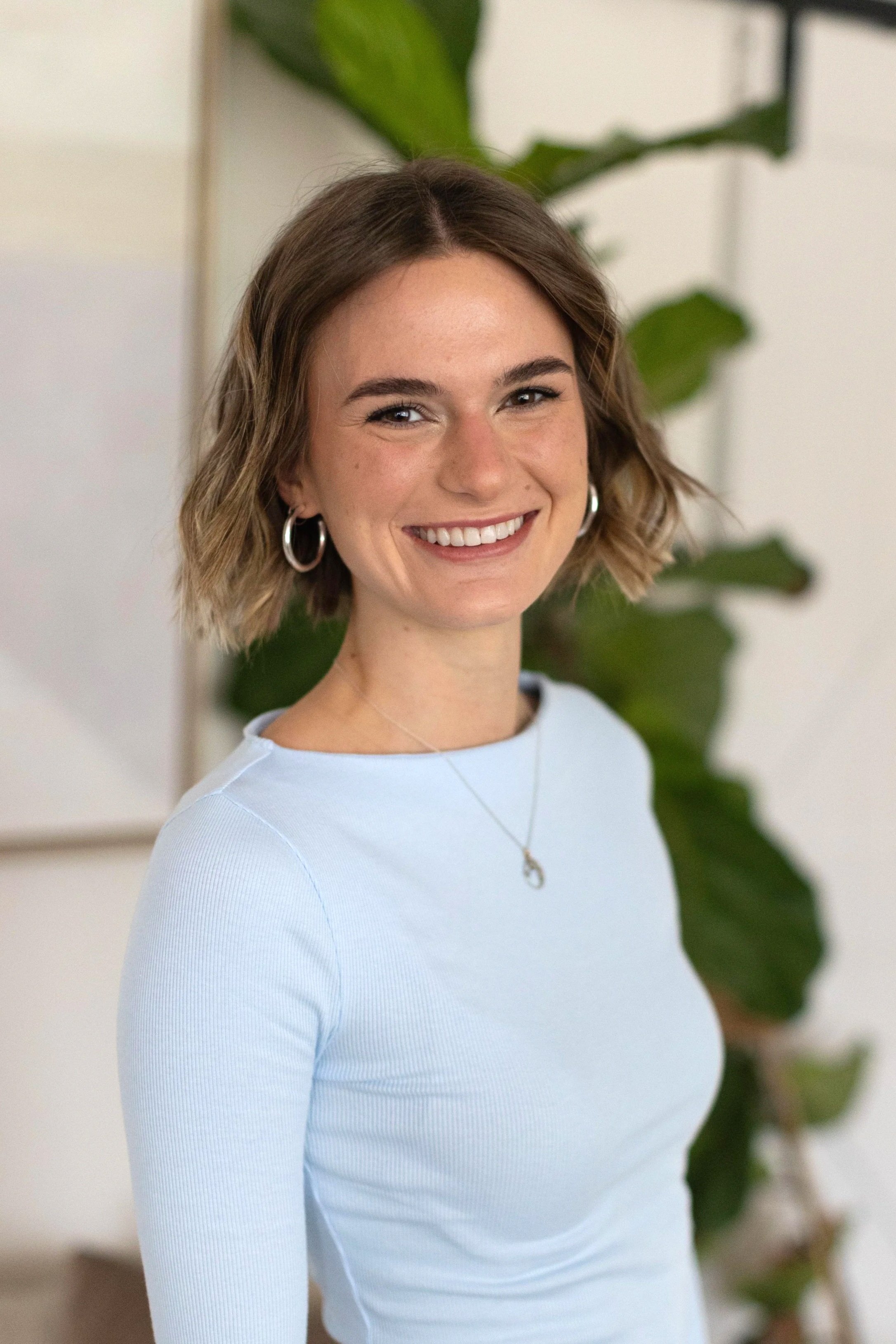 A woman with short, wavy light brown hair smiling and wearing a light blue long-sleeve top, earrings, and a necklace, sitting indoors with a large green plant in the background.