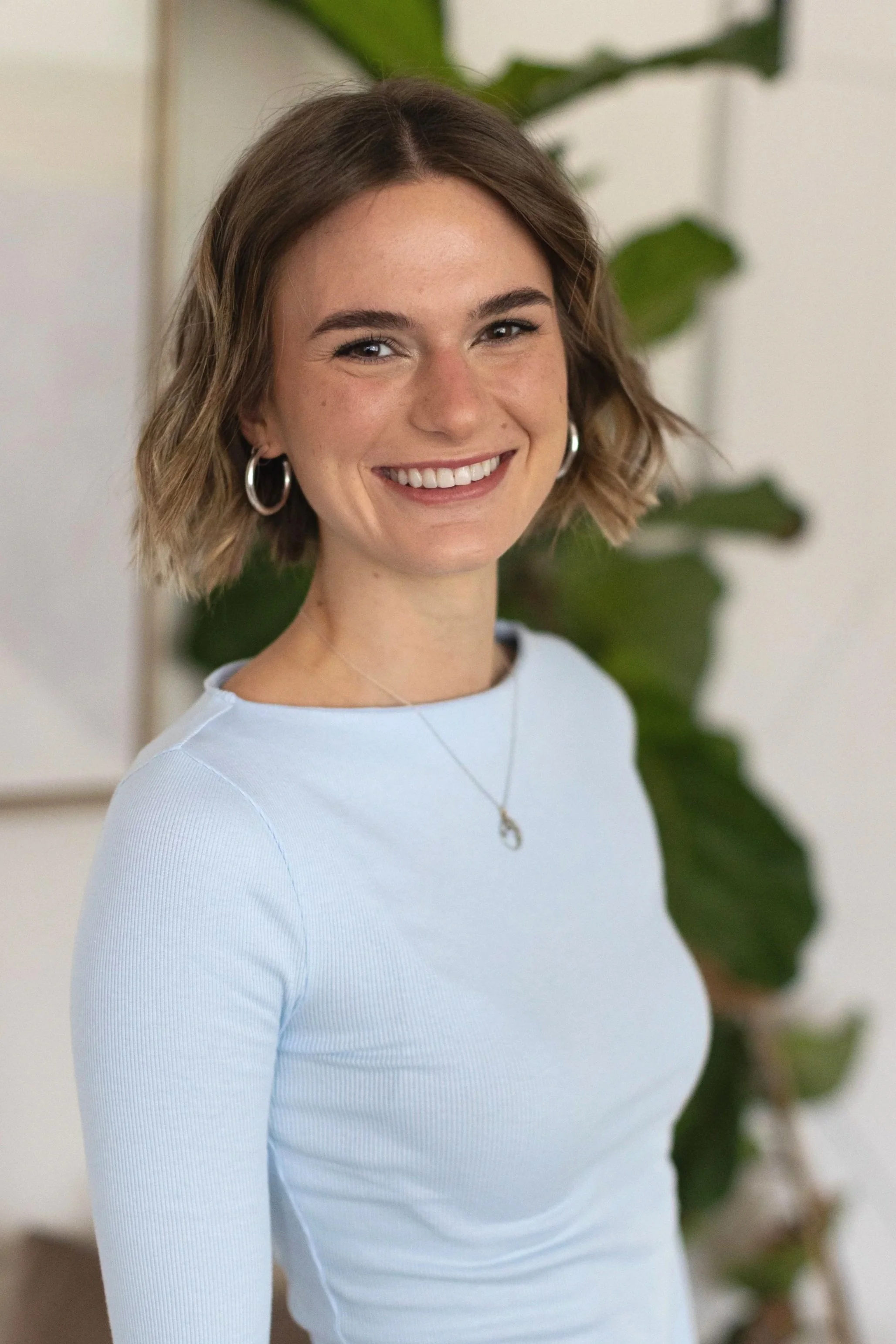 A smiling young woman with short wavy hair wearing a light blue shirt, hoop earrings, and a necklace, sitting indoors with a large green plant in the background.