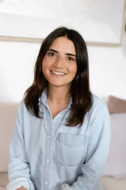 A woman with shoulder-length dark hair smiling, wearing a light blue denim shirt, sitting in a room with a light-colored wall and a beige couch in the background.