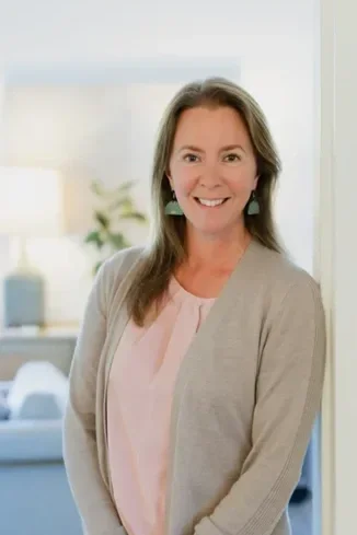 A smiling woman with shoulder-length brown hair, wearing a light pink top and beige cardigan, standing indoors with a window and houseplants in the background.