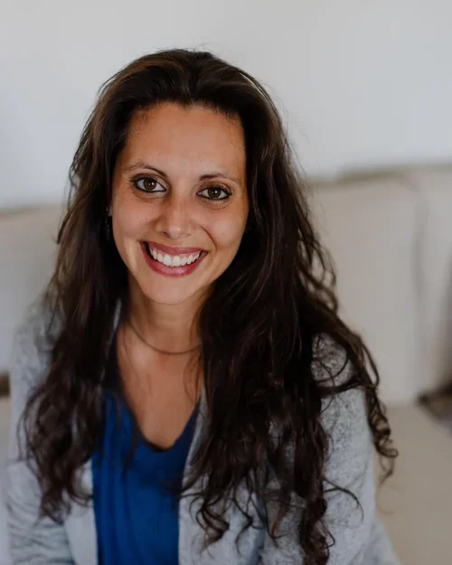 Smiling woman with long wavy brown hair, wearing a blue top and gray cardigan, sitting on a light-colored couch in a bright room.