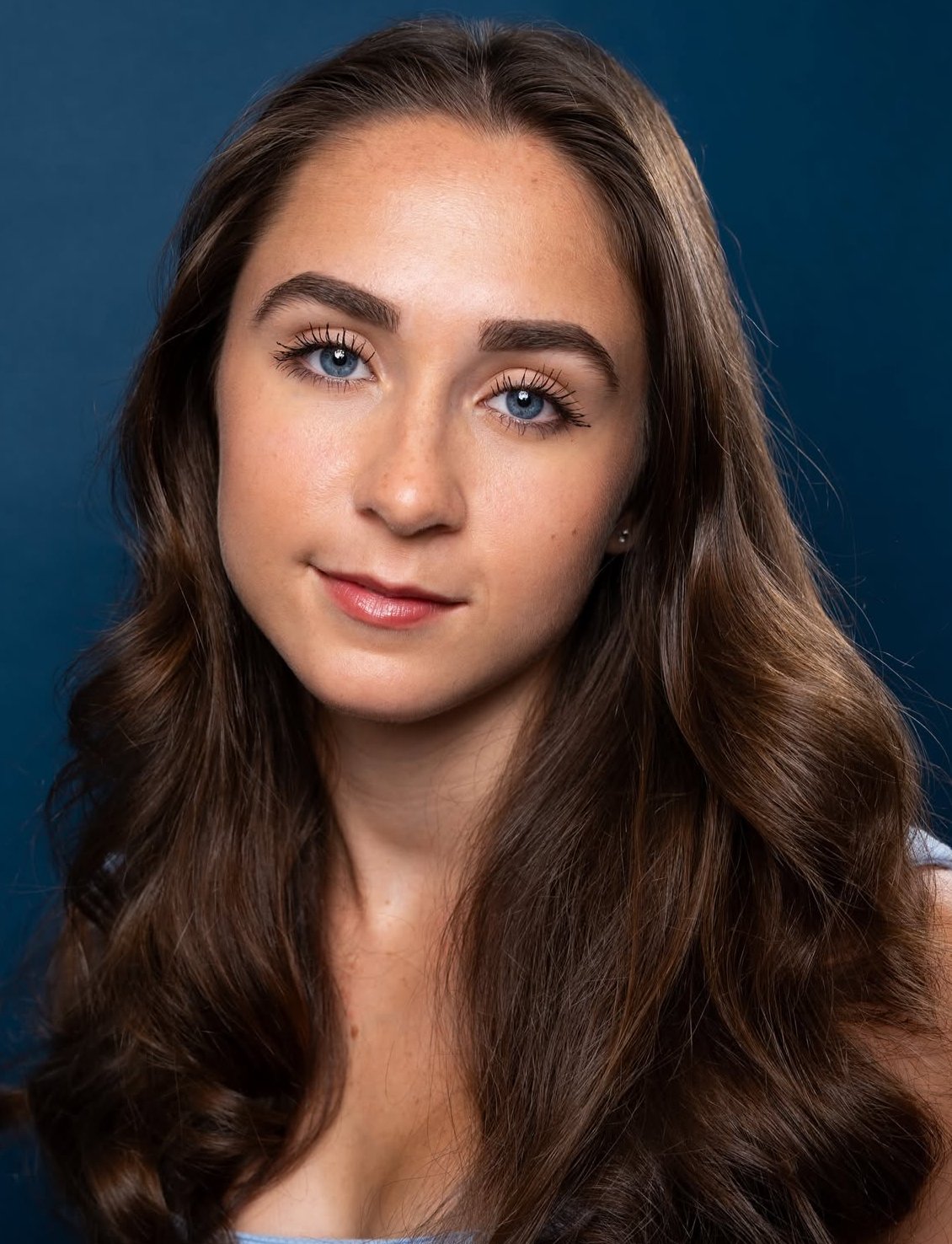 Close-up of a young woman with long, wavy brown hair, blue eyes, and light makeup, smiling slightly against a dark blue background.