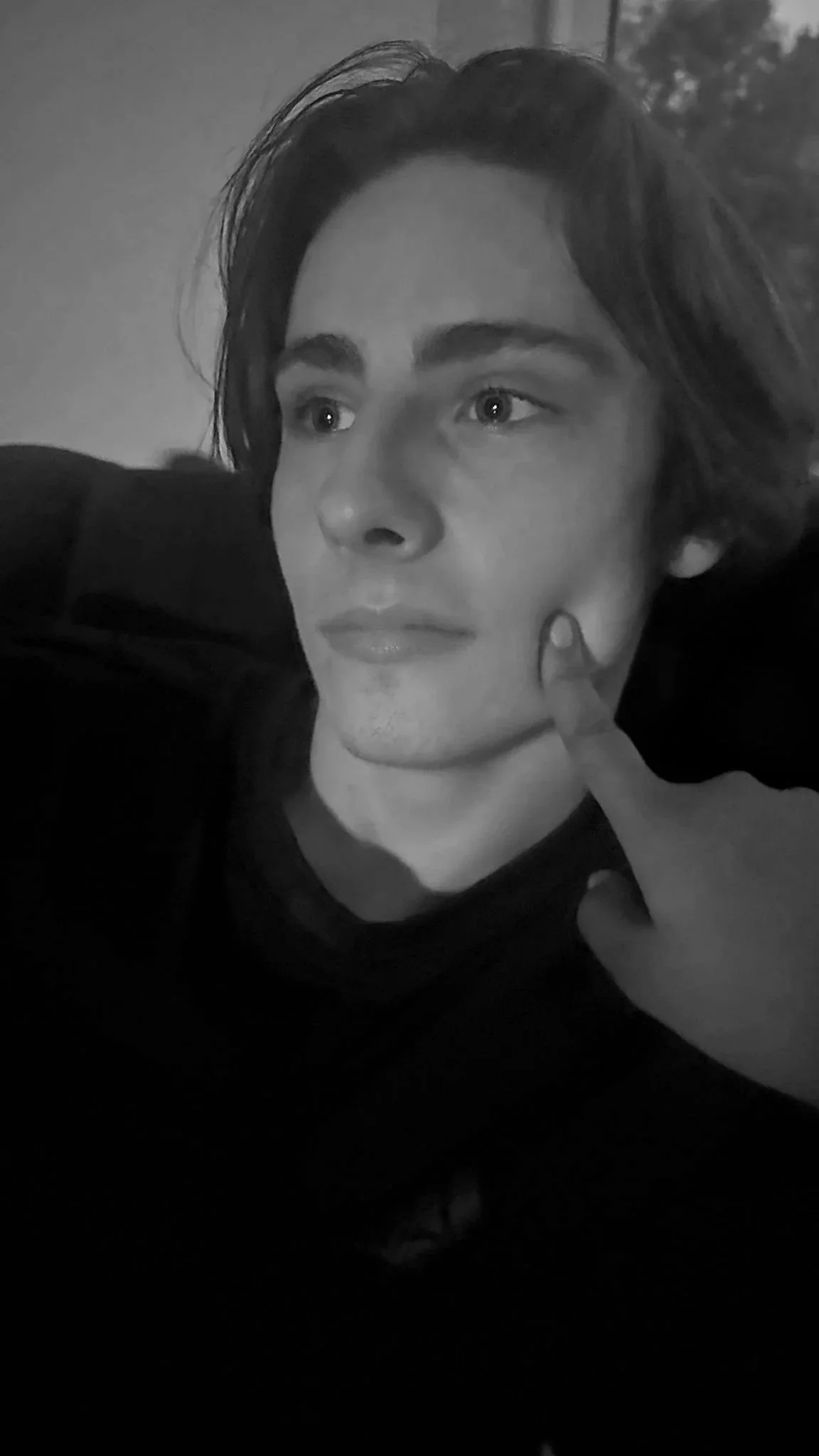 Monochrome photograph of a young man with wavy hair, sitting on a couch with a relaxed posture, looking to the left with a contemplative expression and a finger touching his cheek.