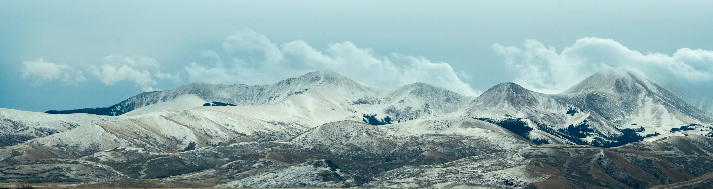 Snow-capped mountains under a cloudy sky with patches of brown and green terrain in the foreground.