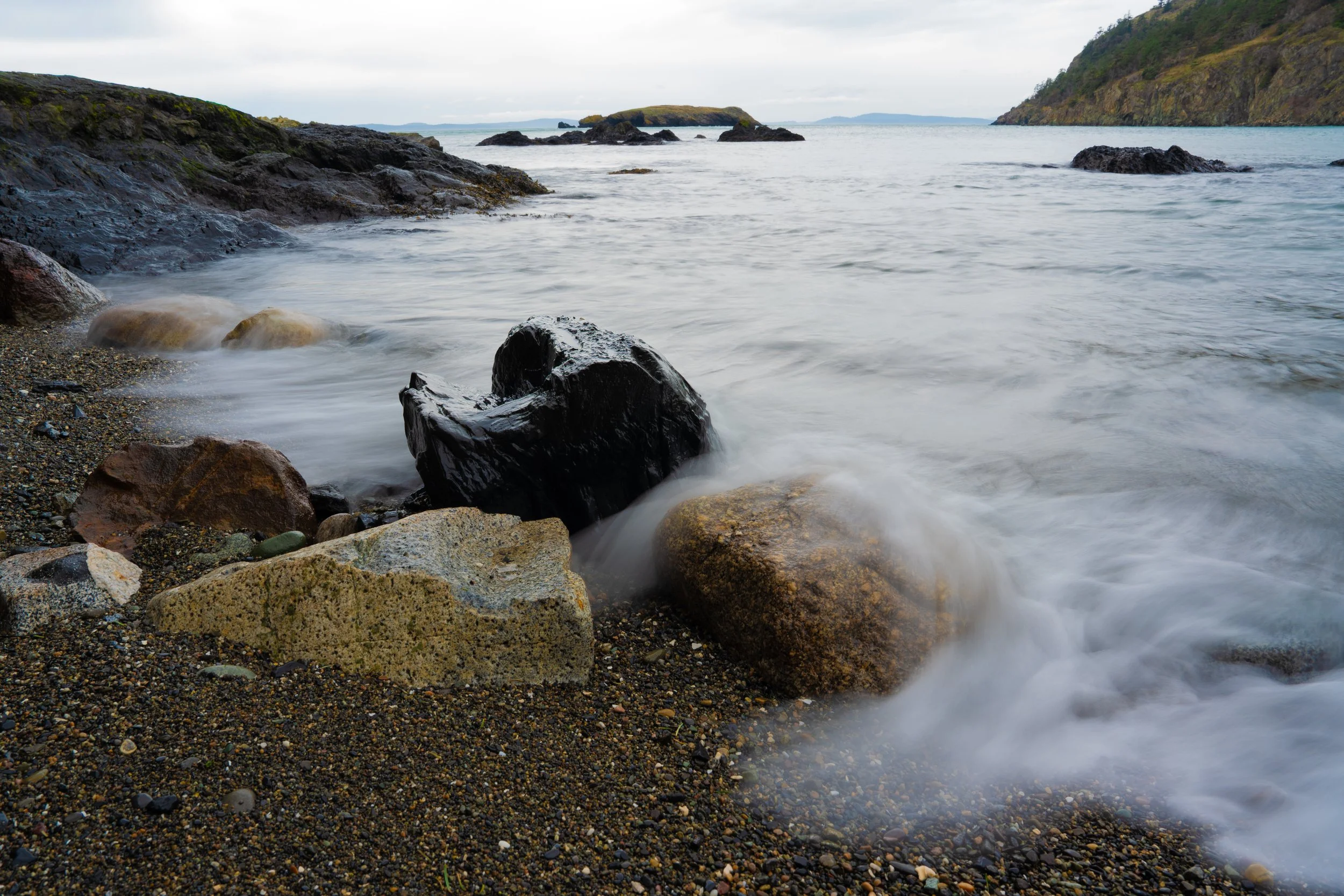 Rocky beach with water flowing over rocks, surrounded by hills and distant islands over a cloudy sky.