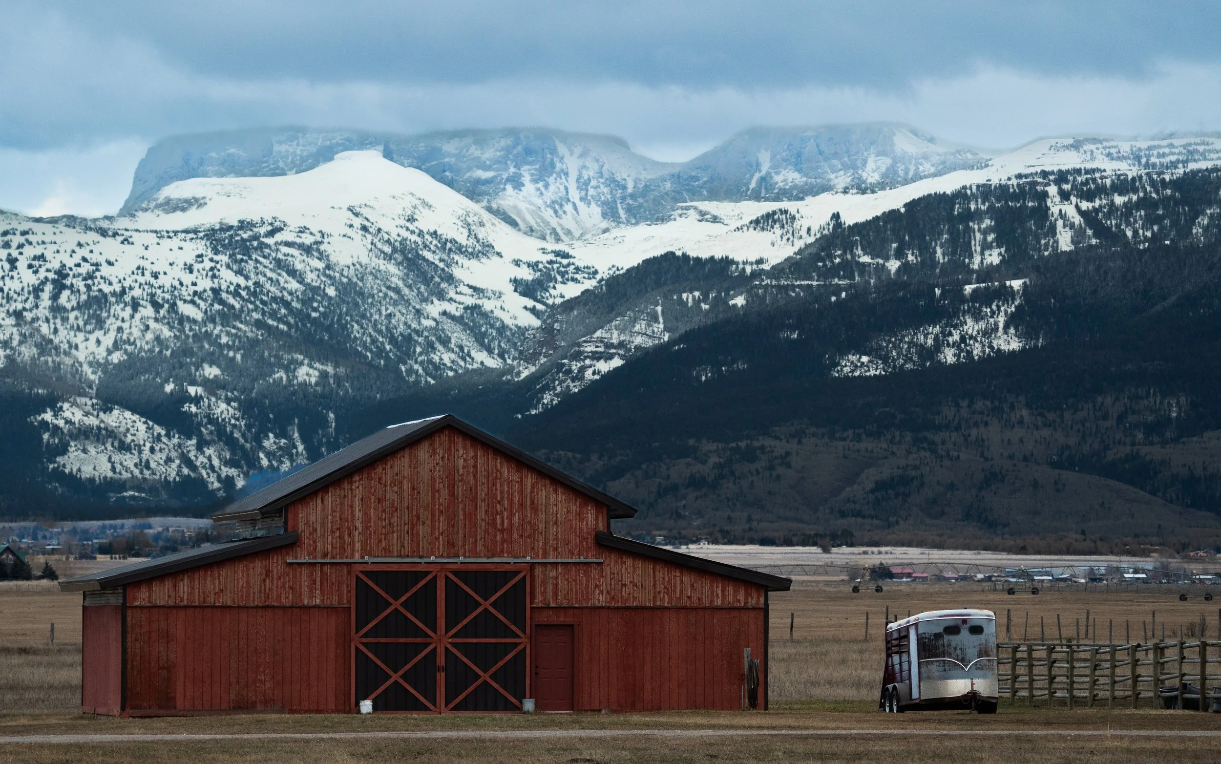 A red barn with a black roof in a rural field, with mountains snow-covered in the background and a white trailer parked nearby.