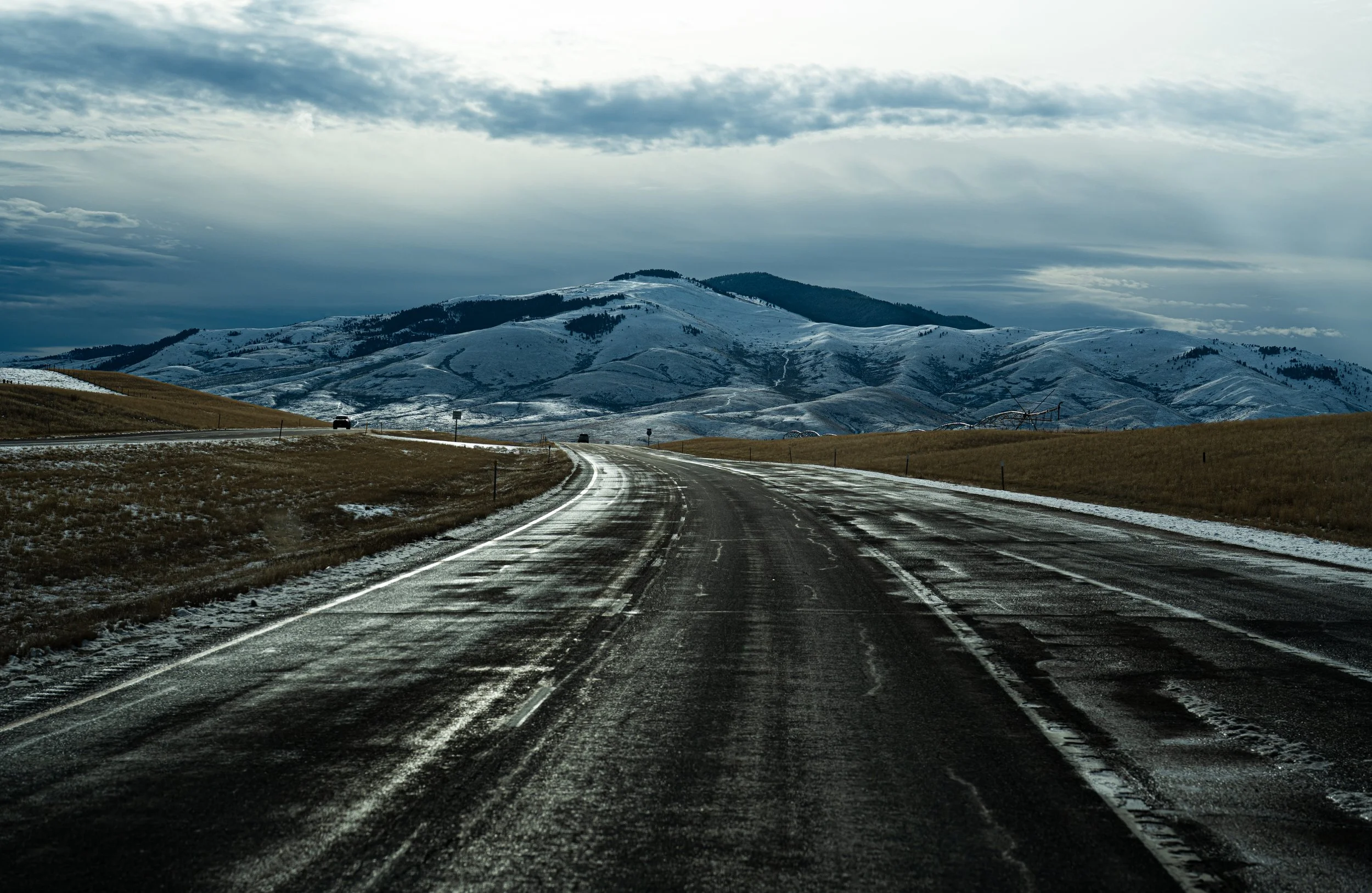 A winding empty road through a mountain landscape with snow-capped hills and cloudy sky.