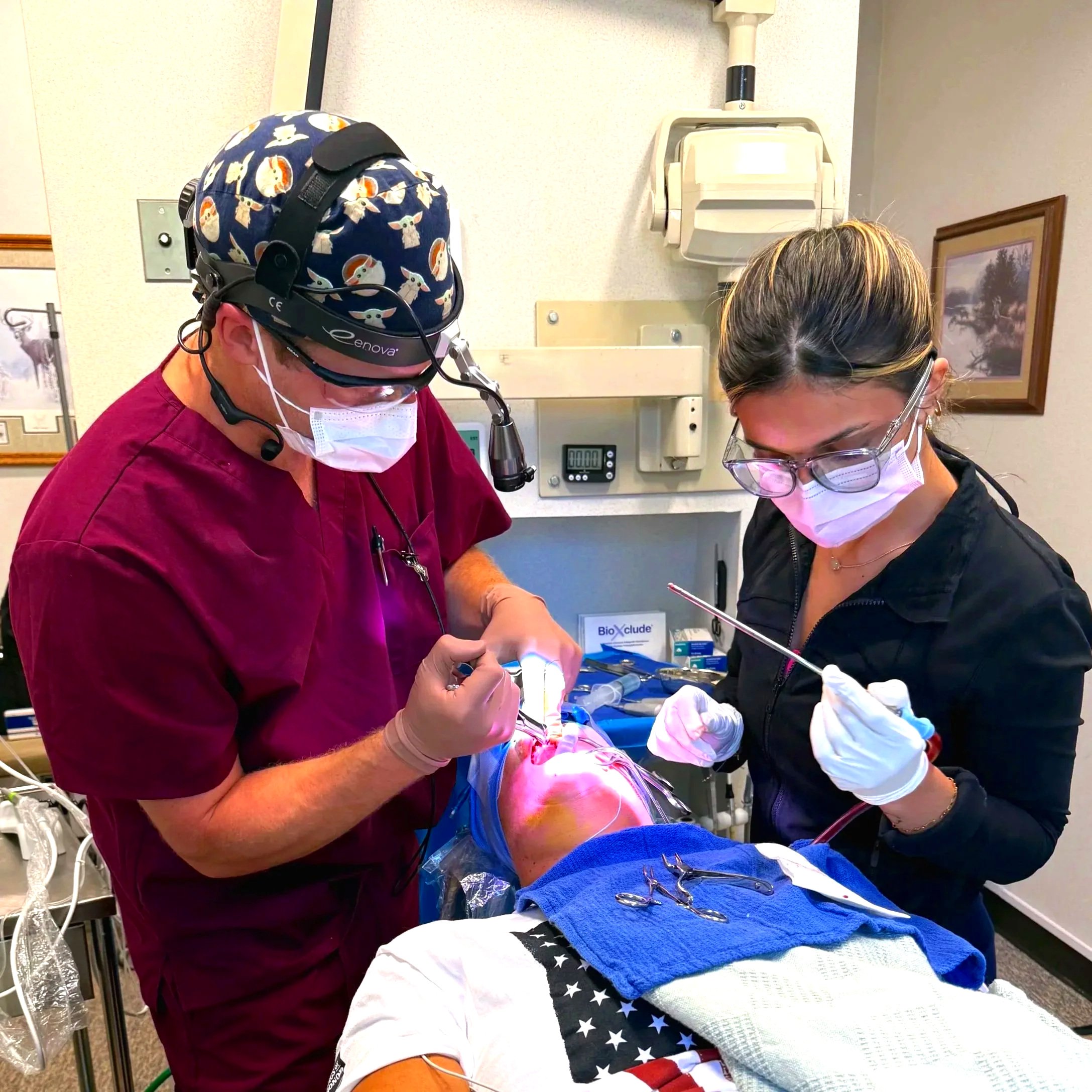 Dentists performing oral surgery on a patient in a dental office while the patient is under general anesthesia and intubated, both the surgeon and assistant are wearing masks and gloves while using surgical dental instruments.