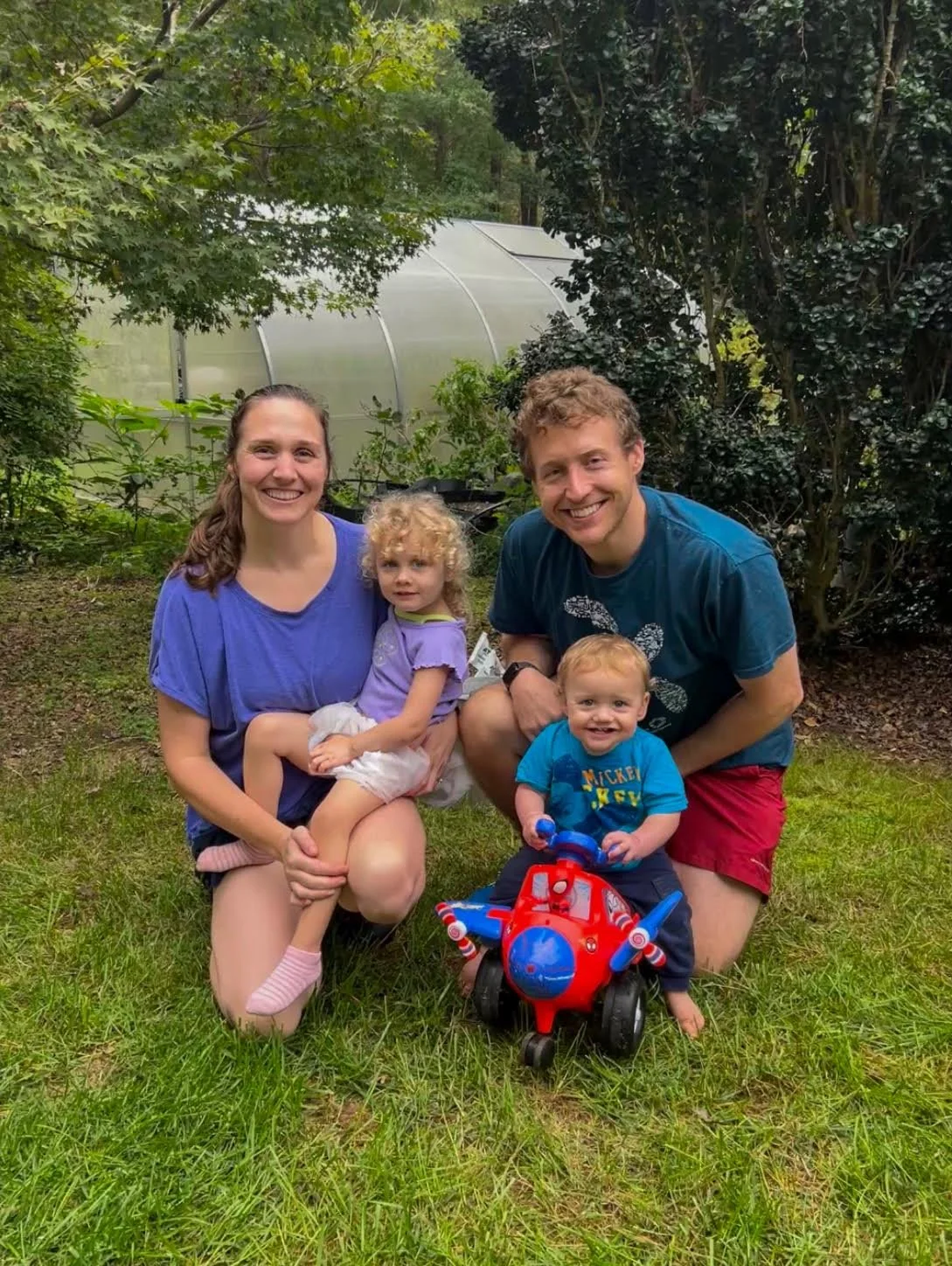 A family of four, two adults and two young children, outdoors on a grassy area with trees and a greenhouse in the background, smiling and posing for a photo. The children are sitting on a colorful toy airplane.