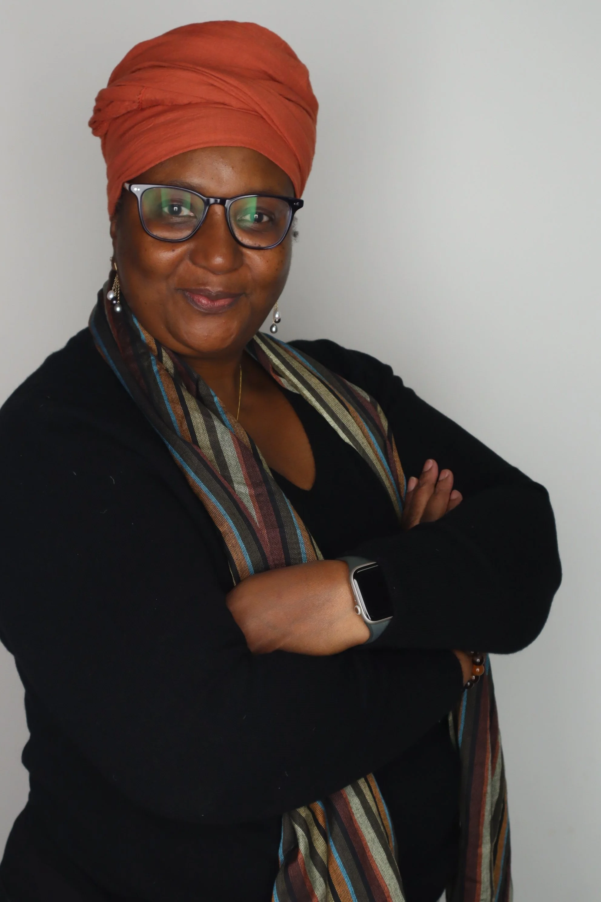 A woman wearing glasses, a burnt orange headwrap, a black top, and a multicolored striped scarf, standing with arms crossed against a plain white wall.