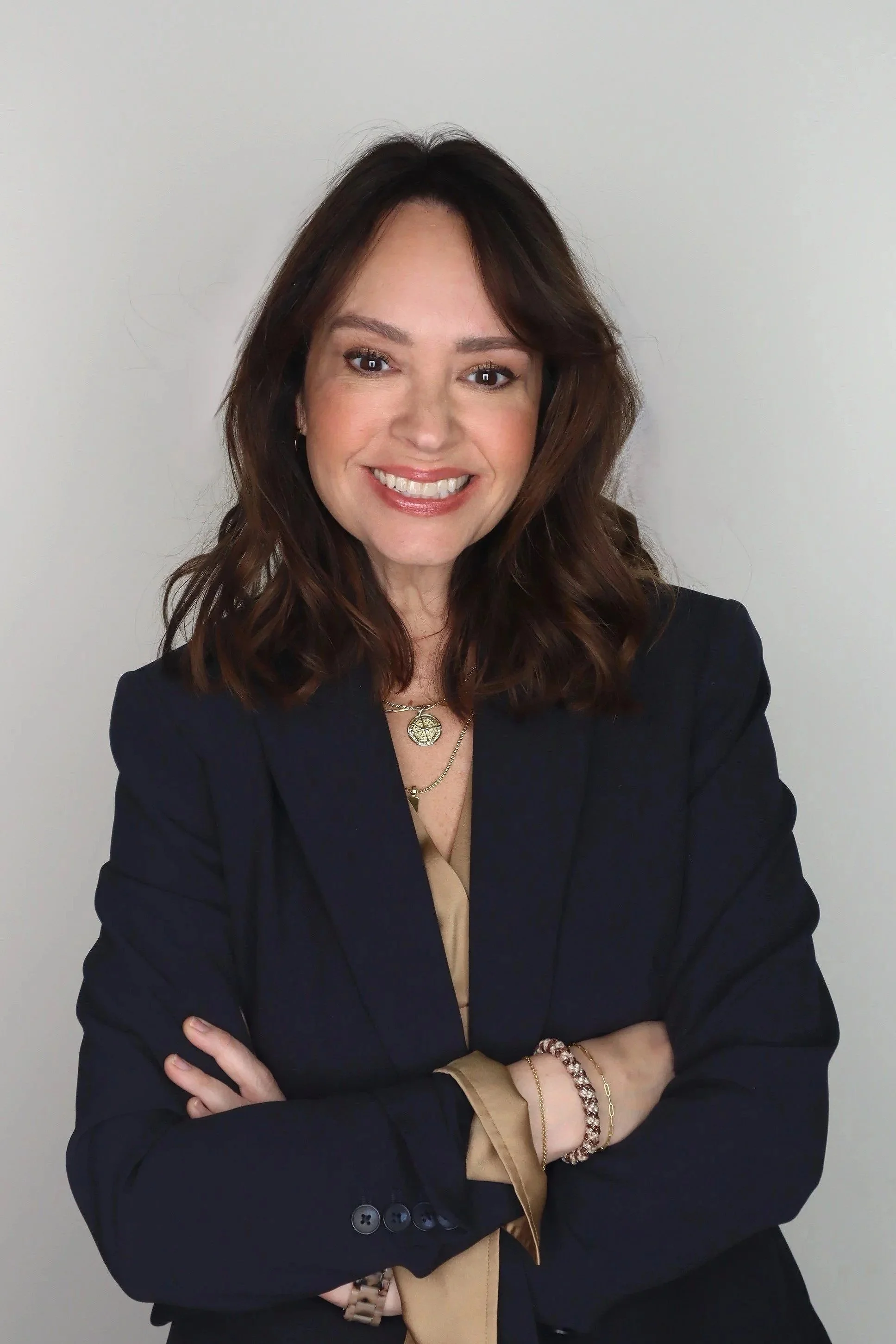 A woman with shoulder-length brown hair smiling, wearing a navy blazer, beige blouse, and layered necklaces, standing against a plain light-colored background.