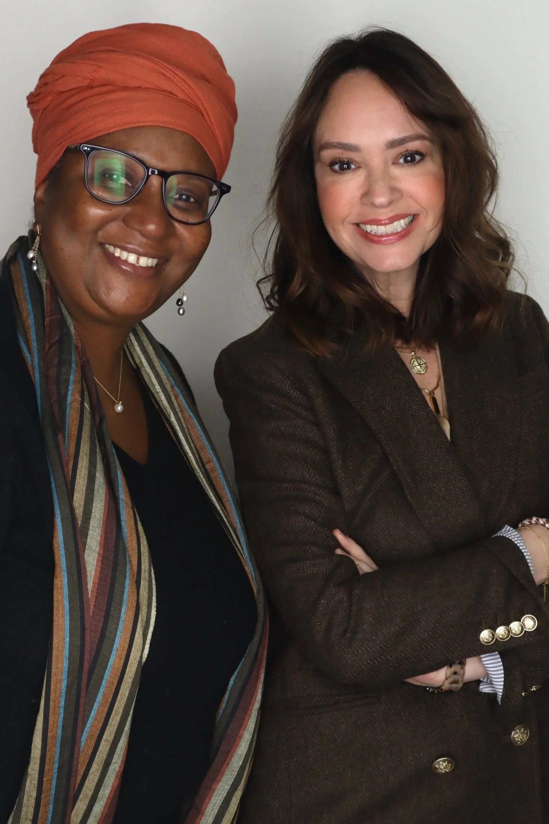 Two women smiling, one wearing glasses and an orange headwrap with a multicolored striped scarf, and the other with brown hair in a dark blazer.
