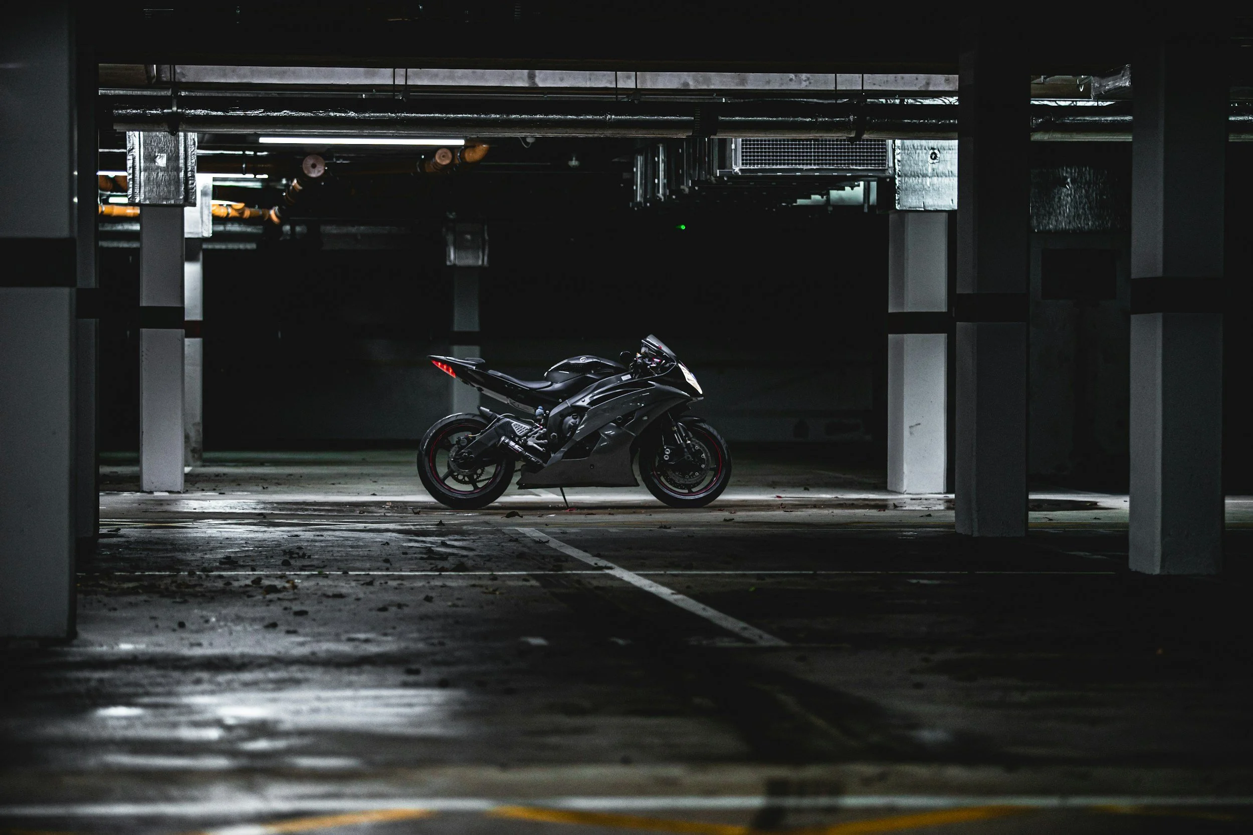 A black motorcycle parked in an underground parking garage with concrete pillars and ceiling pipes, dimly lit.
