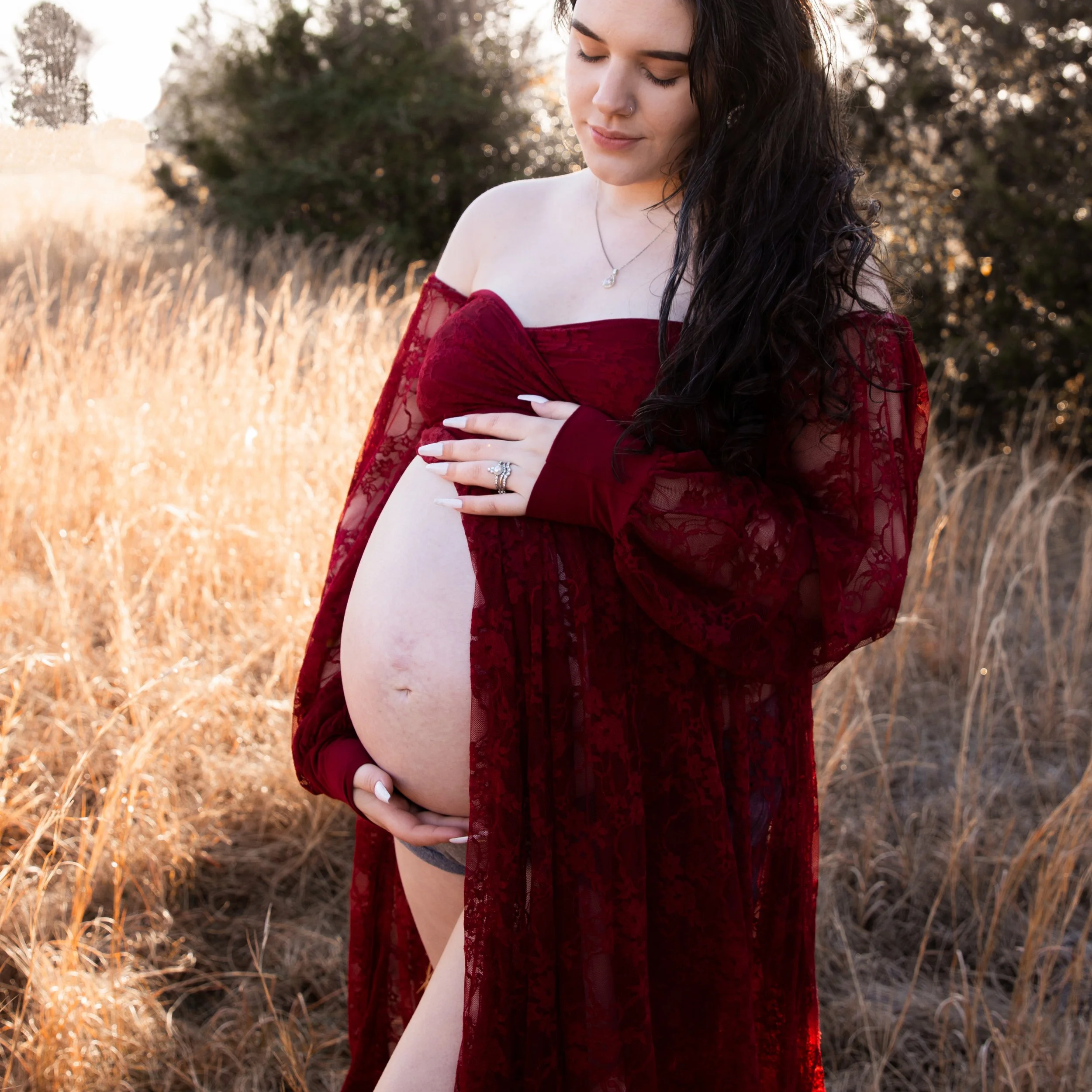 A young woman with long dark curly hair, wearing a red lace off-shoulder dress, standing outdoors in a field with tall golden grass during the late afternoon or early evening. She has her eyes closed and is gently touching her arm, with a serene expression.