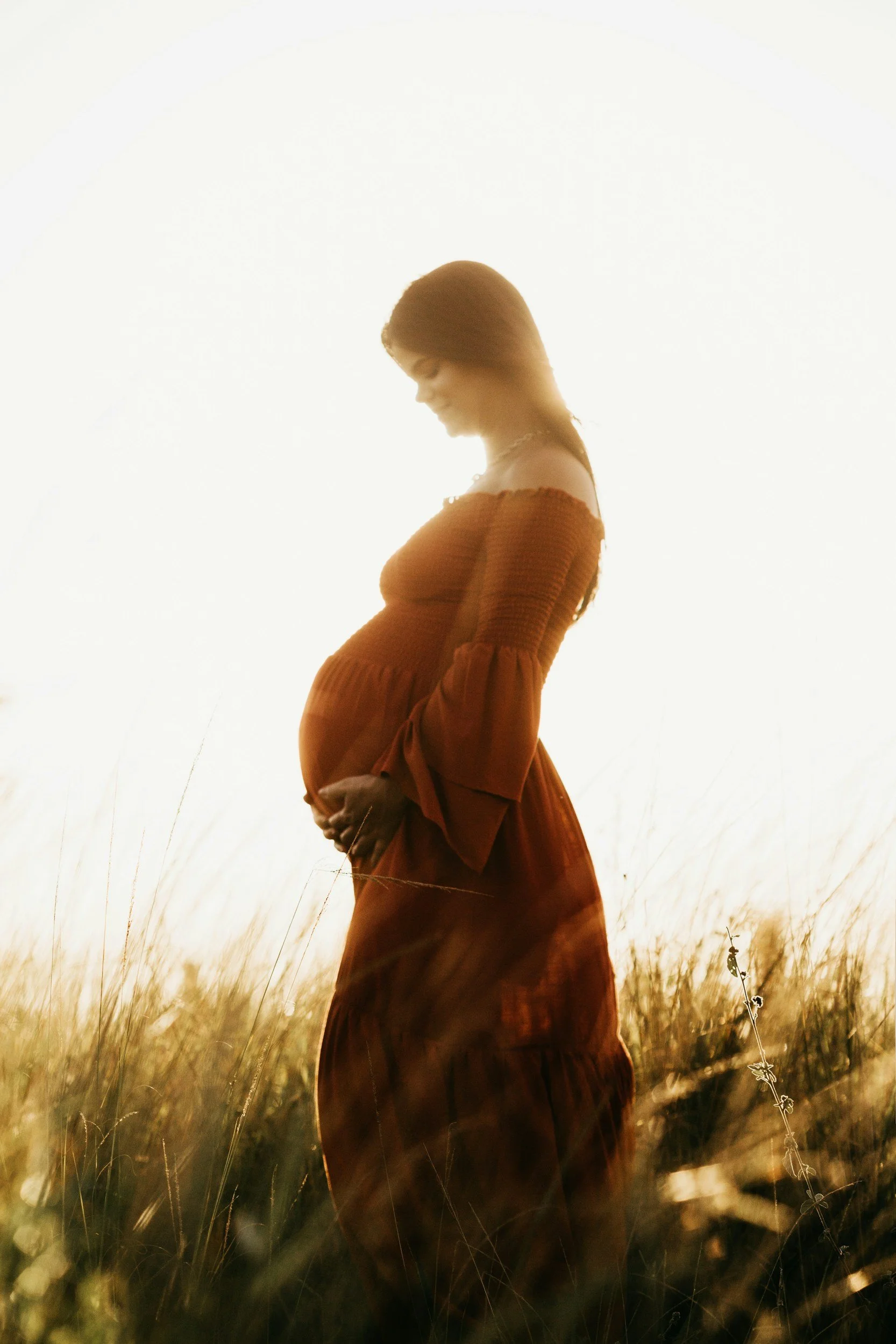 A pregnant woman being supported by light of life birth doula bryan tx in an orange dress stands in a field of tall grass during sunset, gently holding her belly and looking down.